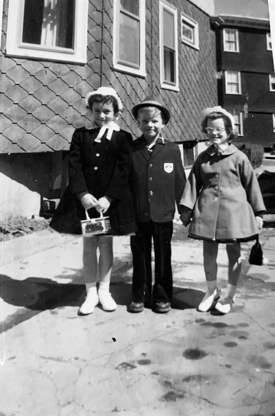 Three children stand happily together in front of a house, dressed in stylish vintage outfits.