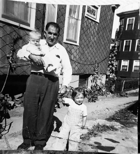 A man stands outside with two children, one in his arms, in a historic neighborhood setting.