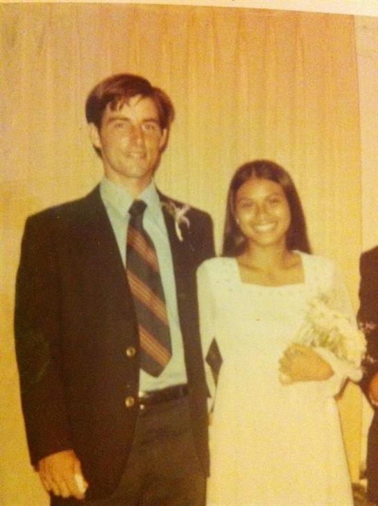 A man and woman smile while wearing formal outfits, holding flowers at a celebration event.