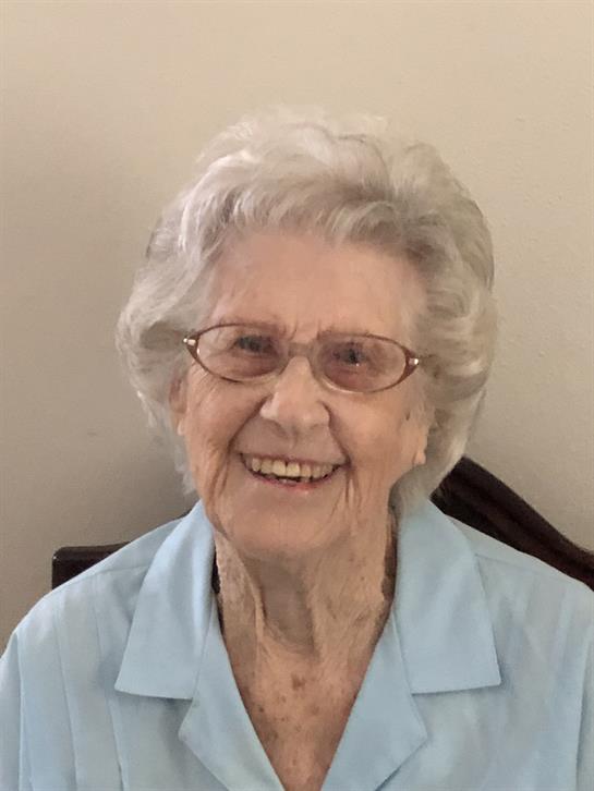 An elderly woman with glasses sits indoors, smiling brightly while wearing a light blue blouse.