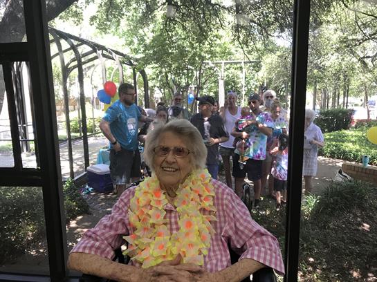 An elderly woman in a pink checked shirt celebrates her birthday with family in a garden.