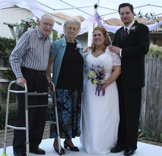 A bride stands proudly with her grandparents and husband in an outdoor celebration.