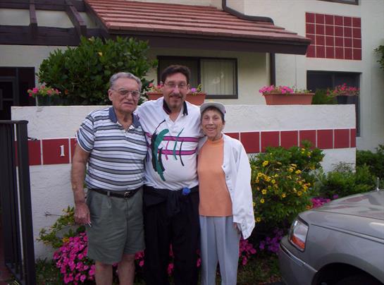 Two men and a woman smile together in front of their home surrounded by colorful flowers.