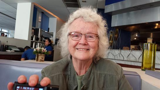 Elderly woman with glasses smiles warmly while taking a picture in a lively cafe setting.