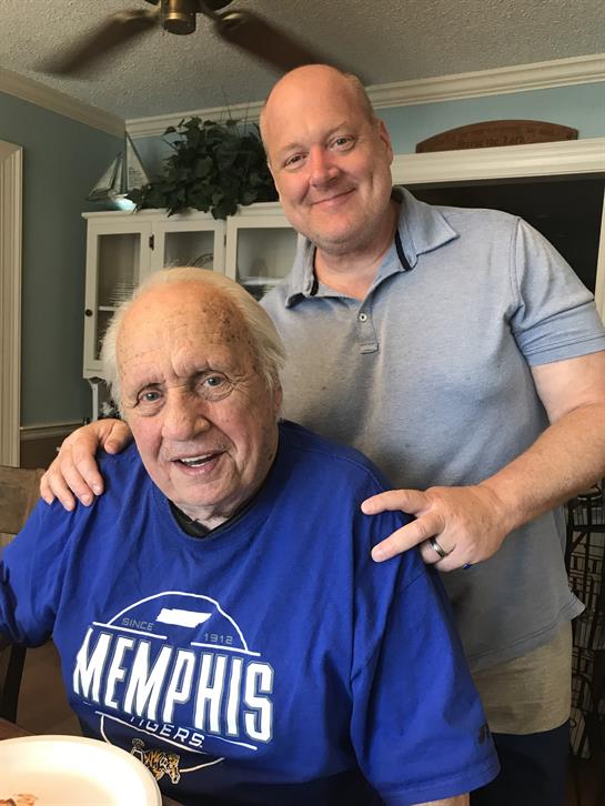 Two men express happiness while posing for a photo in a comfortable living space.