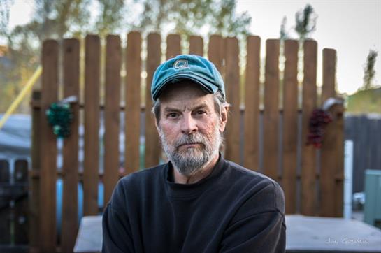 A man with a gray beard and blue cap sits still outside near a wooden fence in the afternoon.