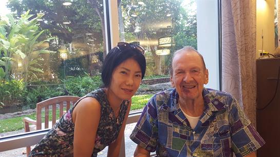Two individuals share a joyful moment at a cafe, surrounded by green scenery and sunlight.