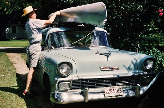 A person works on a vintage car, lifting the hood to inspect the engine under bright sunlight.