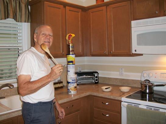 An elderly man stands in a tidy kitchen, tasting food with a wooden spoon while smiling.