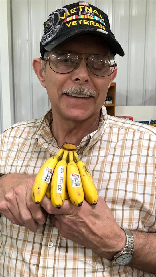 A middle-aged man smiles while presenting four yellow bananas in a market.