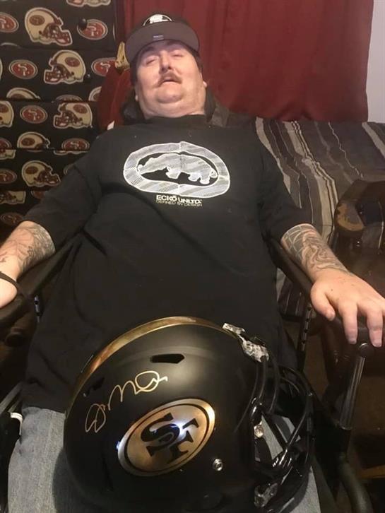 A man sits in a wheelchair at home, wearing a black shirt and surrounded by comfortable decor.