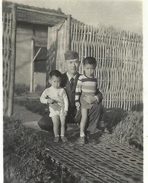 A man crouches beside two young children, both enjoying a sunny day outdoors near a wooden fence.
