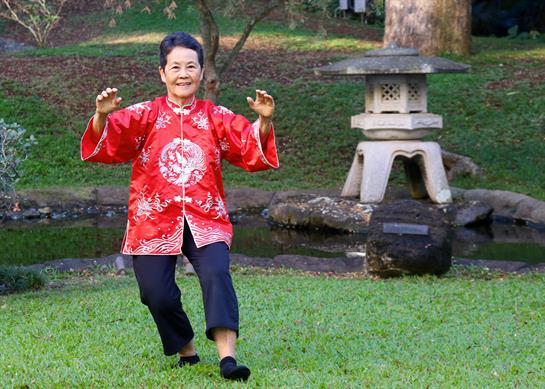 Elderly woman performs tai chi outdoors in a serene park setting while wearing traditional attire.