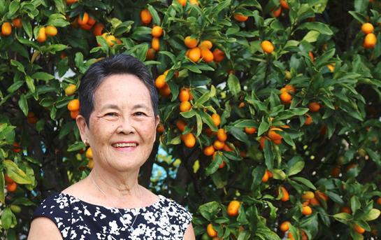 A woman smiles joyfully in a garden filled with lush kumquat trees, showcasing ripe fruits.