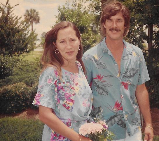 A couple poses with smiles, surrounded by lush greenery on a sunny day during a garden gathering.