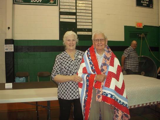 Two seniors stand together smiling warmly, showcasing a vibrant quilt in a community gym setting.