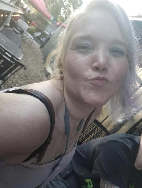 A young woman is making a playful face for a selfie in a busy outdoor cafe filled with people.