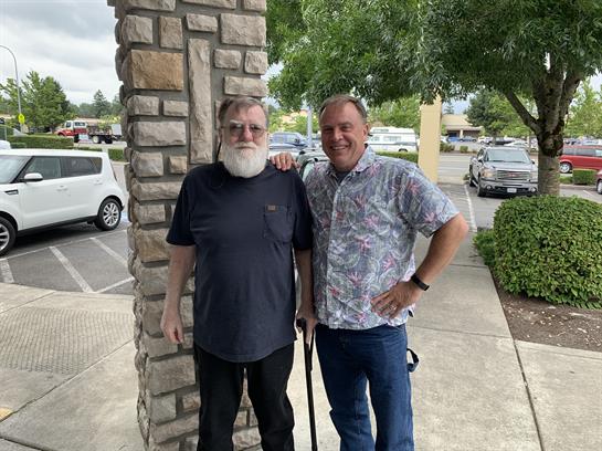 Two men are smiling and posing for a picture outside a stone building near shops on a sunny day.