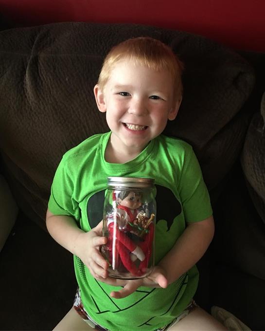 A young boy in a green shirt joyfully displays a jar filled with vibrant toys at home.