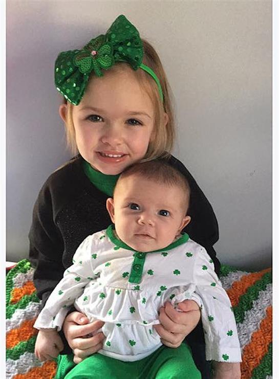 Two young siblings pose happily, dressed in festive green attire, surrounded by warm colors.