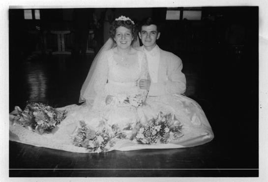 A couple celebrates their wedding, dressed elegantly in white attire, surrounded by flowers.