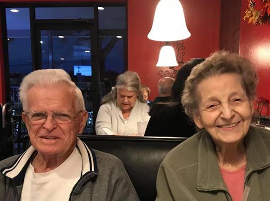 An elderly couple smiles at a restaurant surrounded by diners during the evening.