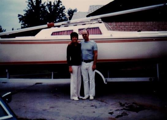 Two people pose together in front of their sailboat at the marina, showcasing their pride and joy.