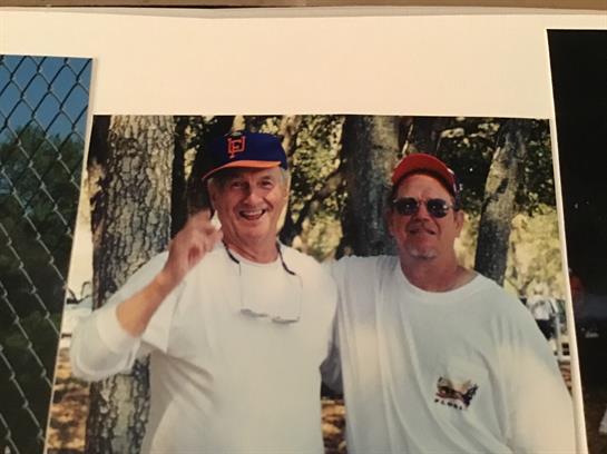 Two men stand together under trees, smiling broadly, enjoying their time outdoors.