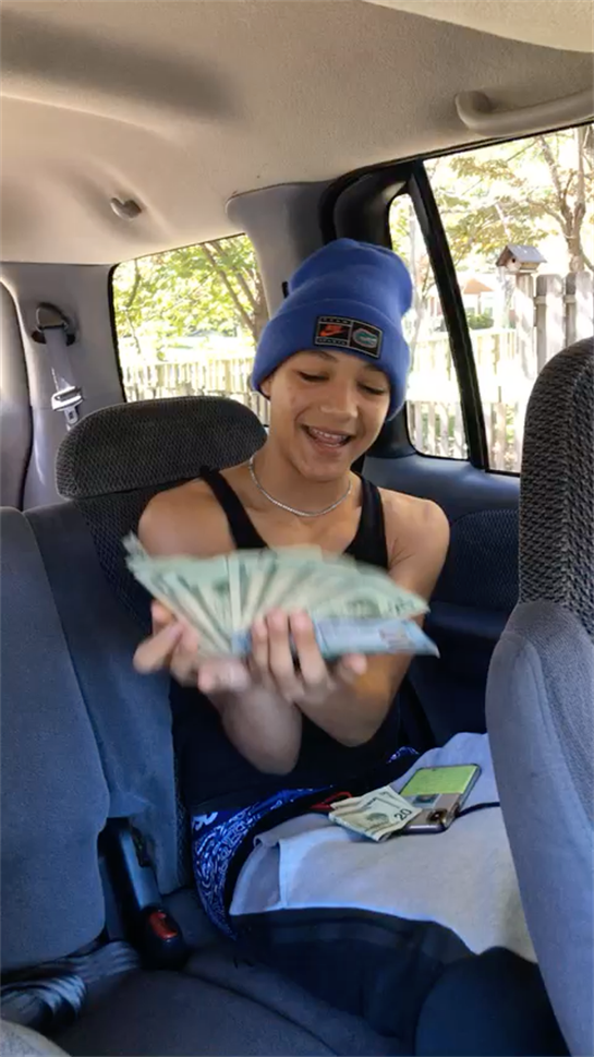 A young boy sits inside a parked vehicle, joyfully displaying a stack of cash while smiling.