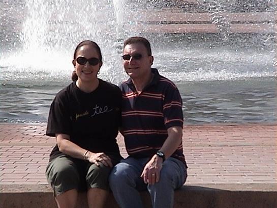 A couple sits together on a warm summer day, smiling by a fountain in a city park.