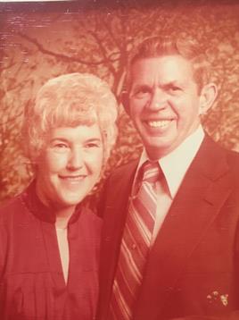 Elderly couple stands closely together in a sunlit outdoor setting, smiling warmly.