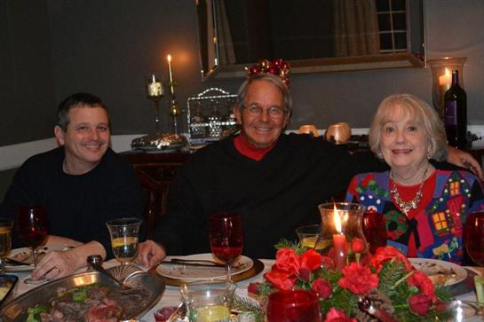 Three people enjoy a festive dinner, surrounded by candles, flowers, and holiday decorations.