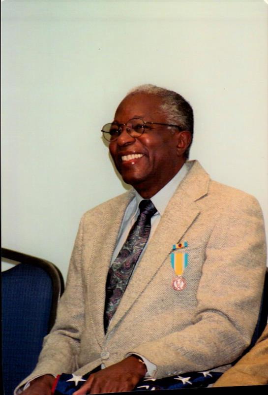 A smiling veteran in formal attire attends a recognition event, showcasing his medals with pride.
