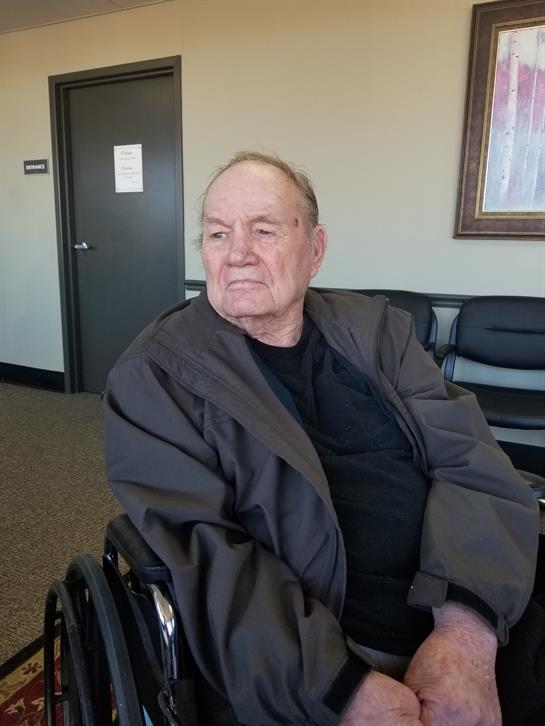 An elderly man in a wheelchair observes his surroundings in a medical facility waiting area.