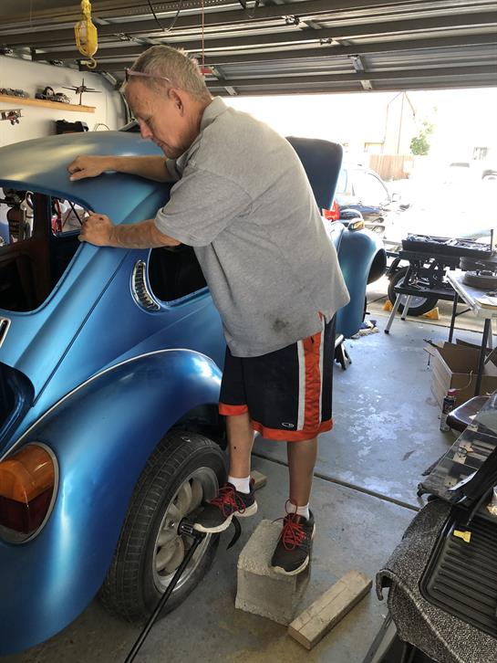 A man works on the back of a blue vintage car in a sunny garage filled with tools.