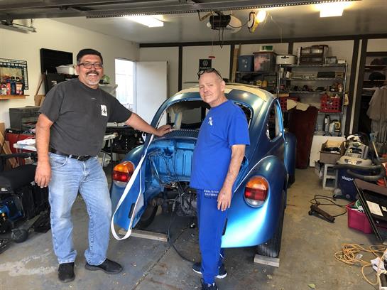Two men stand beside a classic blue car in a garage, showcasing their teamwork and skills.