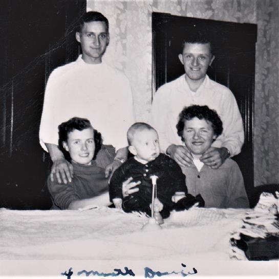 Five family members gather around a table while enjoying a moment with a baby in a warm atmosphere.