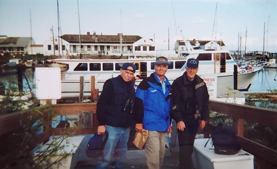 Friends pose with smiles by a boat, surrounded by a marina, enjoying the bright day.
