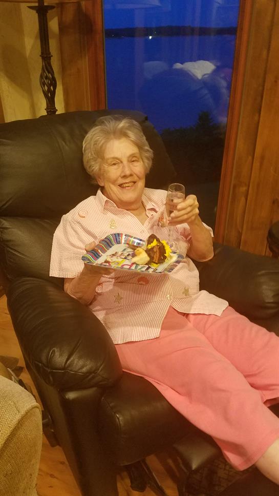 An elderly woman smiles while holding a plate of dessert and a drink in a relaxed home setting.