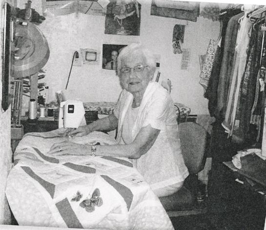 An elderly woman attentively stitches quilts at a sewing table filled with fabric.