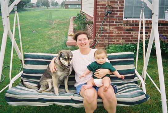 Woman sits on a bench with a child and dog, enjoying a sunny day in a lush backyard.
