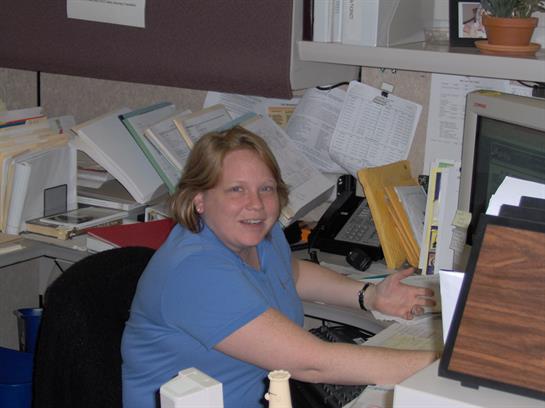 Worker is focused on tasks at a desk filled with documents, office supplies, and equipment.