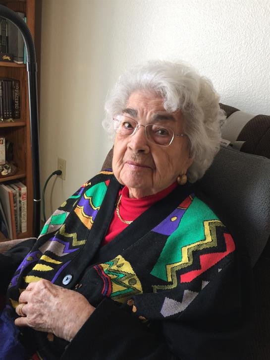 Senior woman with white hair smiles warmly, dressed in a vibrant sweater while seated comfortably.