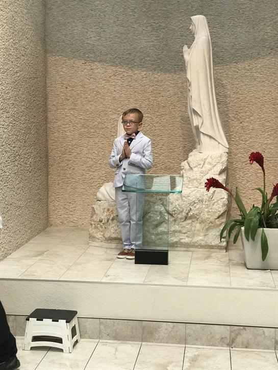 A young boy stands in formal attire in front of a religious statue, engaged in silent prayer.