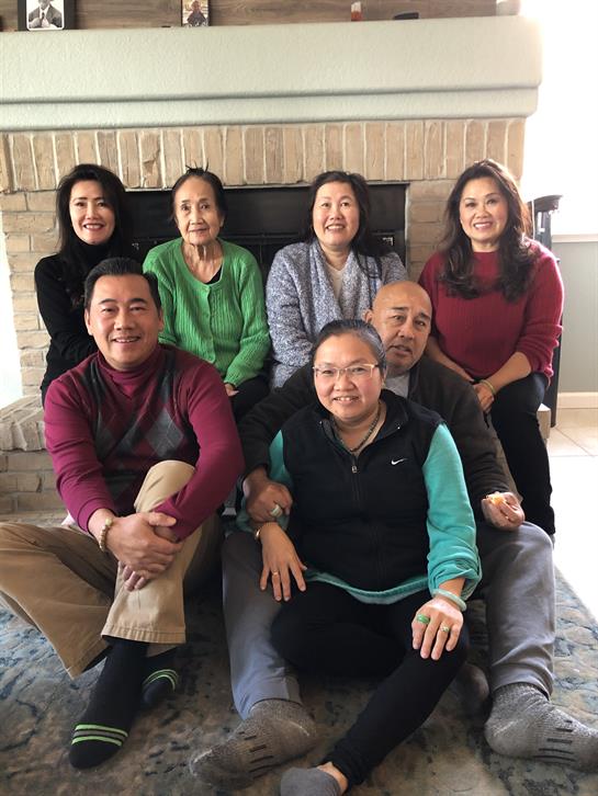 A group of eight friends and family members gather in a cozy living room by the fireplace.