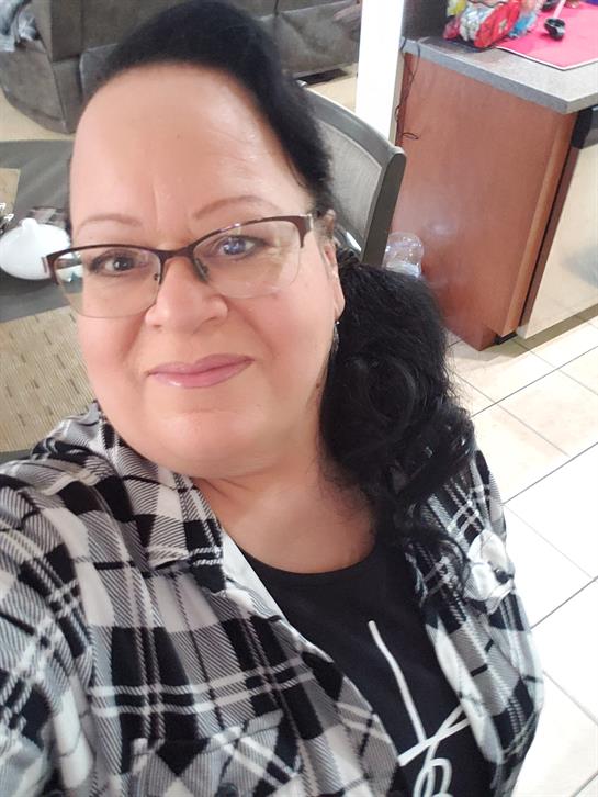 A woman with glasses stands in a kitchen, smiling cheerfully while wearing a plaid shirt.