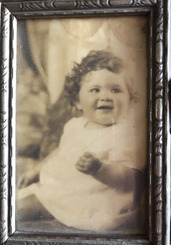 A joyful baby with curly hair is seen wearing a vintage dress and smiling brightly.