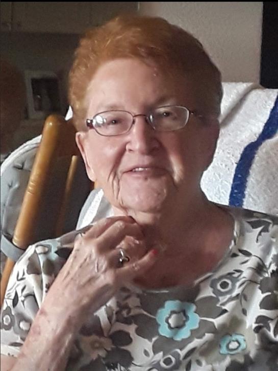 An older woman with glasses smiles warmly while seated in a rocking chair at home.