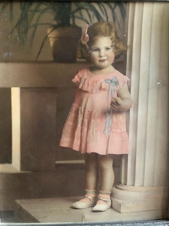 A small girl stands next to a doorway, wearing a pink dress and a hair bow, smiling playfully.