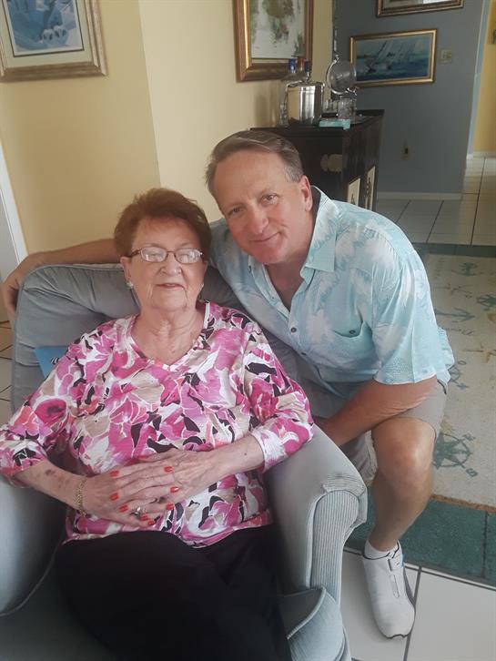 A man sits beside an elderly woman in a light-filled living room, both smiling warmly.